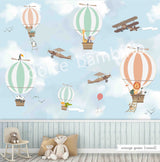 A wall mural featuring various cartoon animals, such as a monkey, penguin, and giraffe, along with hot air balloons and an airplane in a blue sky background. The mural is applied to a wall in a room with a bed and a stuffed bunny toy in the foreground.