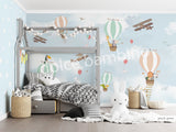 A wall mural featuring various cartoon animals, such as a monkey, penguin, and giraffe, along with hot air balloons and an airplane in a blue sky background. The mural is applied to a wall in a room with a bed and a stuffed bunny toy in the foreground.
