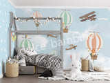 A wall mural featuring various cartoon animals, such as a monkey, penguin, and giraffe, along with hot air balloons and an airplane in a blue sky background. The mural is applied to a wall in a room with a bed and a stuffed bunny toy in the foreground.