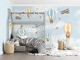 A wall mural featuring various cartoon animals, such as a monkey, penguin, and giraffe, along with hot air balloons and an airplane in a blue sky background. The mural is applied to a wall in a room with a bed and a stuffed bunny toy in the foreground.