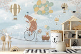 A wall mural featuring a cartoon bear riding a bicycle with a hot air balloon and various other playful animals, on a baby blue background.