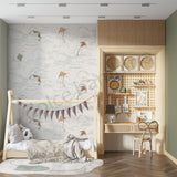 A styled children's bedroom featuring a light wood bed frame, a built-in wooden desk and shelving unit with toys and decor, and a wall covered in a soft gray wallpaper with a kite pattern. A light wood chair sits in front of the desk, and a round rug is on the floor.