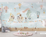 A whimsical wall mural featuring a bear on a bicycle with a wagon full of various cartoon animals, set against a pink background with hot air balloons and a cloudy sky.