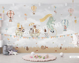 A whimsical wall mural featuring a bear on a bicycle with a wagon full of various cartoon animals, set against a grey background with hot air balloons and a cloudy sky.