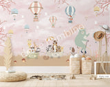 Large nursery wall mural in soft pink and beige tones featuring hot air balloons floating among birds and cherry blossom branches. A mint-green bear rides a vintage bicycle while woodland animals travel in a wagon below, creating a whimsical storybook scene for a baby's room