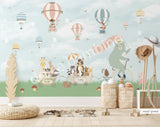 Large nursery wall mural in mint green and dark green tones featuring hot air balloons floating among birds and cherry blossom branches. A mint-green bear rides a vintage bicycle while woodland animals travel in a wagon below, creating a whimsical storybook scene for a baby's room