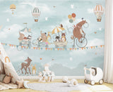 A children's wall mural featuring an illustration of a bear riding a bicycle with various animals such as a hedgehog, raccoon, elephant, panda, and fox in a wagon, against a sky filled with hot air balloons and birds.