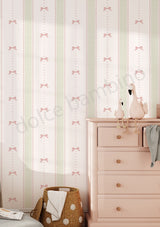 A styled nursery room corner featuring light pink and sage green wallpaper with a repeating bow pattern, a pale pink wooden dresser, a woven basket, and decorative swan figurines.