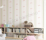 A styled children's room corner featuring a wall covered in vertical striped wallpaper with pink bow motifs, a light wood bookshelf filled with books and storage bins, and a folded knit blanket on top.