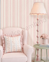 A styled interior scene featuring pink and white vertically striped wallpaper with a small gold dot pattern, a pale pink armchair with a matching striped pillow, a pink floor lamp, and a small round side table with flowers.