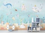 A room with 'Under the Sea' themed wallpaper featuring illustrations of sea creatures such as an octopus, whale, sea horses, and starfish in pastel colors. The room is styled with sea-themed bedding, a sofa, and decorative items.
