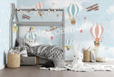 A wall mural featuring various cartoon animals, such as a monkey, penguin, and giraffe, along with hot air balloons and an airplane in a blue sky background. The mural is applied to a wall in a room with a bed and a stuffed bunny toy in the foreground.