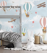 A wall mural featuring various cartoon animals, such as a monkey, penguin, and giraffe, along with hot air balloons and an airplane in a blue sky background. The mural is applied to a wall in a room with a bed and a stuffed bunny toy in the foreground.