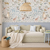 A styled children's daybed in a nursery or playroom setting, featuring a safari-themed animal wallpaper with giraffes, elephants, lions, and trees. The room includes a light blue paneled wainscoting, a woven straw pendant light, a small side table, and a basket of toys.