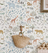 A styled interior scene showing a wall covered in whimsical safari animal wallpaper with illustrated giraffes, elephants, zebras, lions, birds, and trees in muted pastel and earthy tones. A woven straw basket hangs from a giraffe-shaped hook on the wall, with part of a framed print and a small cabinet visible in the room setting.