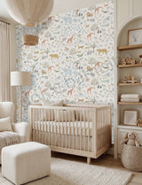 A styled nursery room featuring a large wall mural with a safari animal pattern in soft pastel colors. In front of the wall is a light wood crib, with a cream upholstered armchair, ottoman, floor lamp, and built-in shelving on the right.