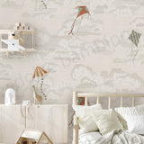 A styled children's bedroom featuring a wall covered in a beige and gray cloud-patterned wallpaper with illustrated kites. A light wood bed with white pillows is in the foreground, along with a white sideboard, shelf, and toys.