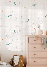 A styled children's room corner featuring wallpaper with a white grid background and playful cartoon children, birds, and geometric motifs in muted teal, orange, and yellow. A pale pink wooden dresser stands on the right, with a woven basket and folded white cloth at the bottom left.