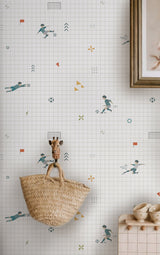 A styled interior scene showing wallpaper with a white grid and scattered playful sports illustrations, a woven straw basket hanging from a giraffe-shaped hook, and part of a wooden frame and shelf visible.