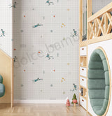 A children's bedroom corner featuring wallpaper with a white grid background and scattered playful illustrations of children, soccer balls, and small geometric shapes. The room includes light wood furniture, a green circular bench, and a small rug.