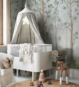 A nursery room with a crib, white furniture, and a wall mural featuring green birch trees.