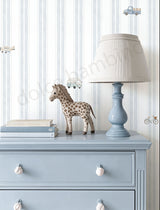 A styled nursery corner featuring a light blue wooden dresser, a matching blue table lamp with a cream shade, a giraffe figurine, and stacked books against a white and blue striped wallpaper background.