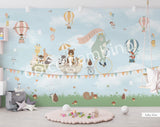 A whimsical wall mural featuring a bear on a bicycle with a wagon full of various cartoon animals, set against a grey background with hot air balloons and a cloudy sky.