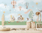 Large nursery wall mural in baby blue and dark green tones featuring hot air balloons floating among birds and cherry blossom branches. A mint-green bear rides a vintage bicycle while woodland animals travel in a wagon below, creating a whimsical storybook scene for a baby's room