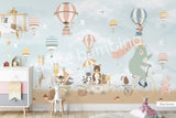 Large nursery wall mural in blue brown and beige tones featuring hot air balloons floating among birds and cherry blossom branches. A mint-green bear rides a vintage bicycle while woodland animals travel in a wagon below, creating a whimsical storybook scene for a baby's room