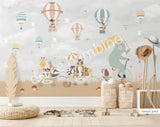 Large nursery wall mural in grey and beige tones featuring hot air balloons floating among birds and cherry blossom branches. A mint-green bear rides a vintage bicycle while woodland animals travel in a wagon below, creating a whimsical storybook scene for a baby's room
