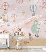 Large nursery wall mural in soft pink and beige tones featuring hot air balloons floating among birds and cherry blossom branches. A mint-green bear rides a vintage bicycle while woodland animals travel in a wagon below, creating a whimsical storybook scene for a baby's room
