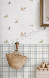 A styled interior wall featuring a white wallpaper with a playful pattern of small animal illustrations and tiny orange dots, paired with a pale green and beige plaid lower band. A woven straw tote bag hangs from a giraffe-shaped hook on the wall.