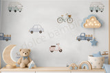 A children's bedroom wall decorated with pastel-colored toy car and bicycle wall decals, featuring a teddy bear, wooden moon lamp, and storage basket on a light wood shelf.
